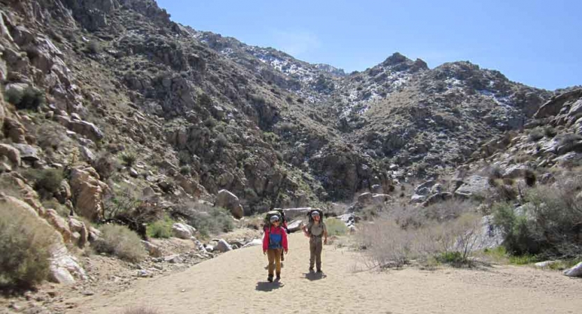 Two backpackers stand on a sandy trail framed by rocky hills